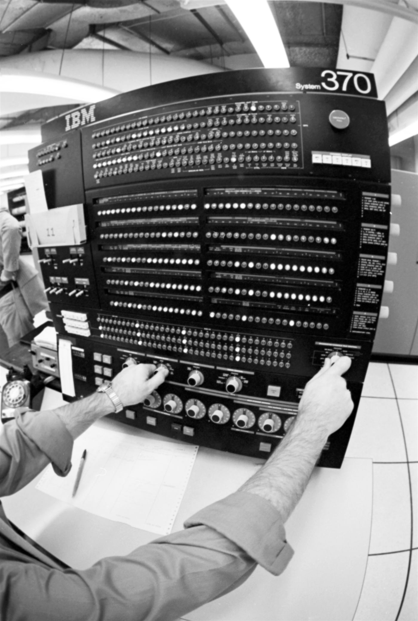 An operator's hands on the front panel of an IBM System/370 console, covered in indicator lamps and rotary dials.