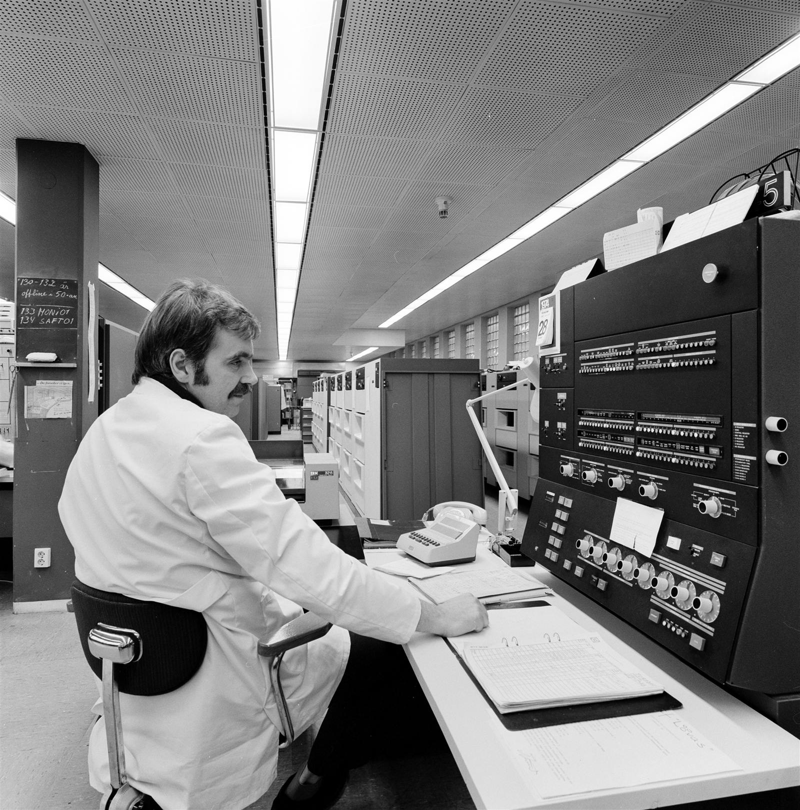 A computer operator in a white coat at the console of an IBM System/370 installation in the mid-1970s, with rows of mainframe cabinets behind him.