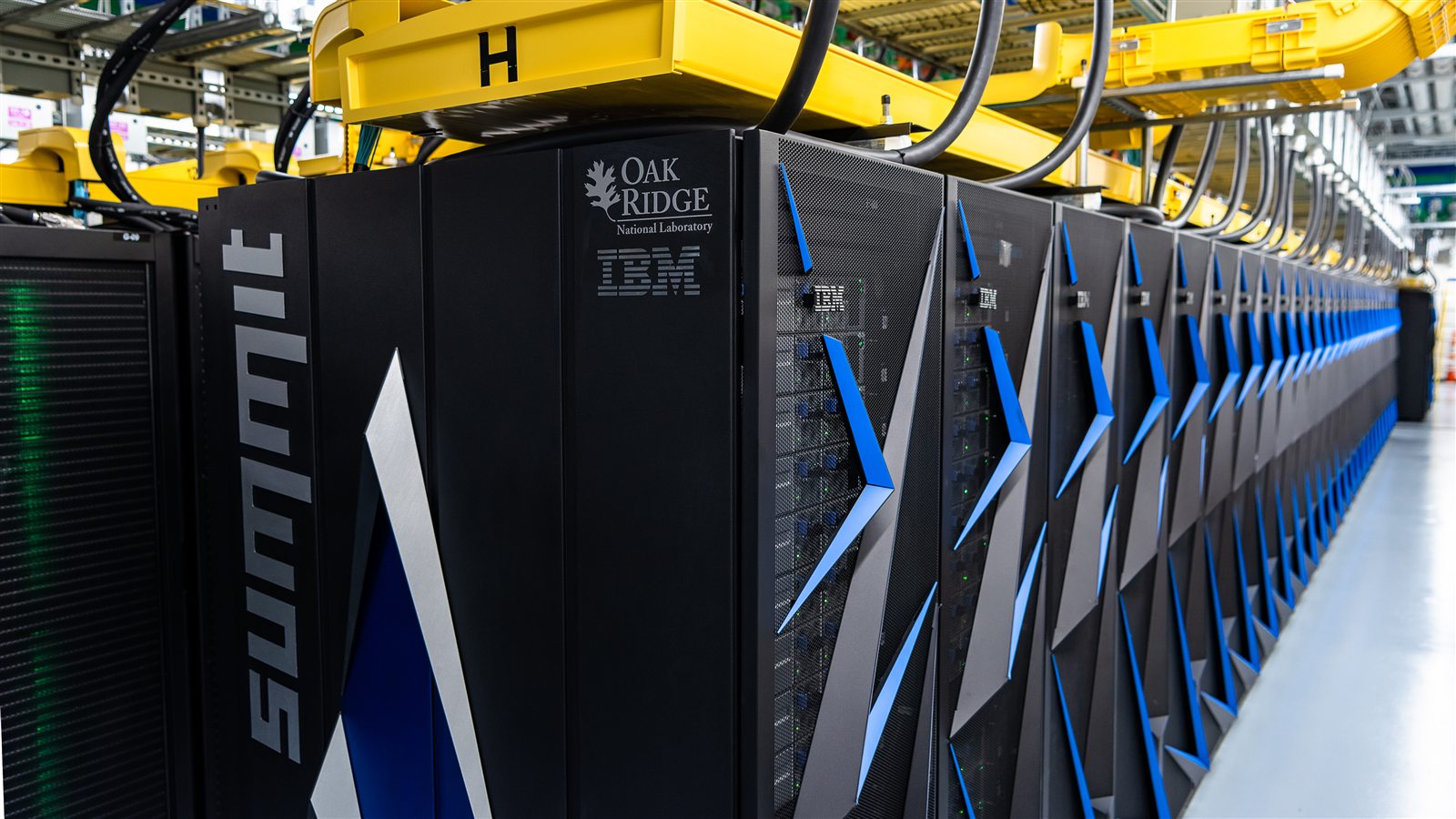 A long row of black supercomputer cabinets under yellow overhead cable trays in a machine hall.